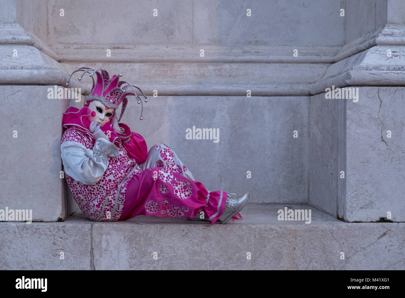 Mujer en rosa Jester's traje, sombrero y máscara, sentada en los escalones de la iglesia San Giorgio Maggiore, Venecia. Foto de stock