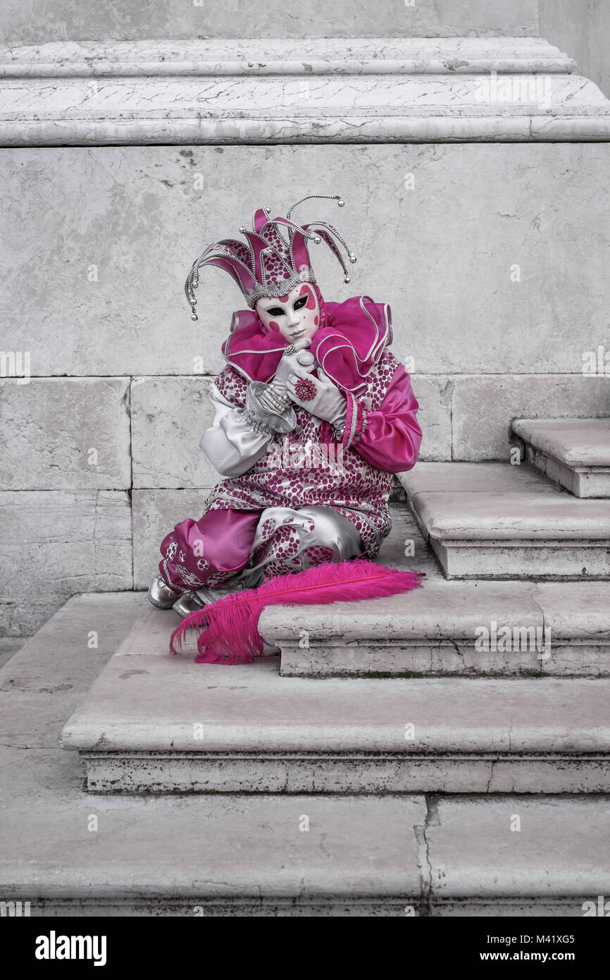 Mujer en rosa Jester's traje, sombrero y máscara, sentada en los escalones de la iglesia San Giorgio Maggiore, Venecia. Foto de stock