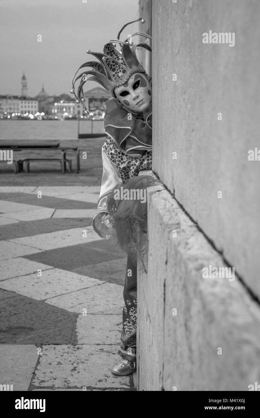 Mujer en el traje de bufón, sombrero y máscara, sentada en los escalones de la iglesia San Giorgio Maggiore, Venecia. Fotografiado en monocromo Foto de stock