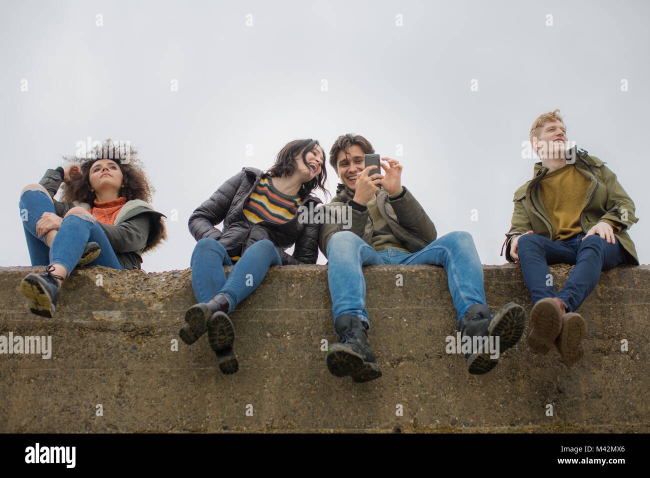 Grupo de jóvenes amigos sentado en una pared mirando un smartphone Foto de stock