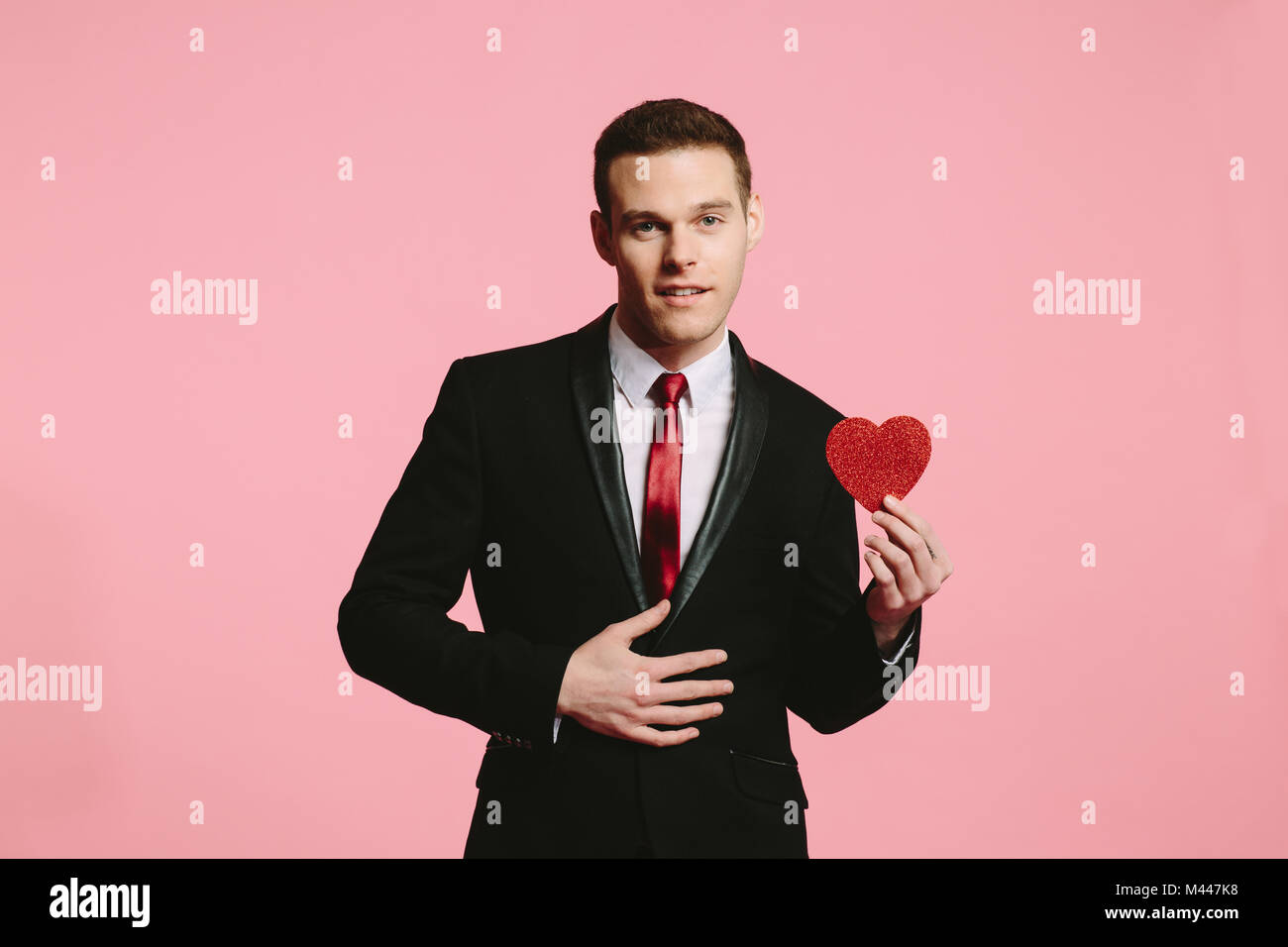 Guapo en un traje negro y corbata roja ofrece un corazón rojo sobre fondo de color rosa Foto de stock