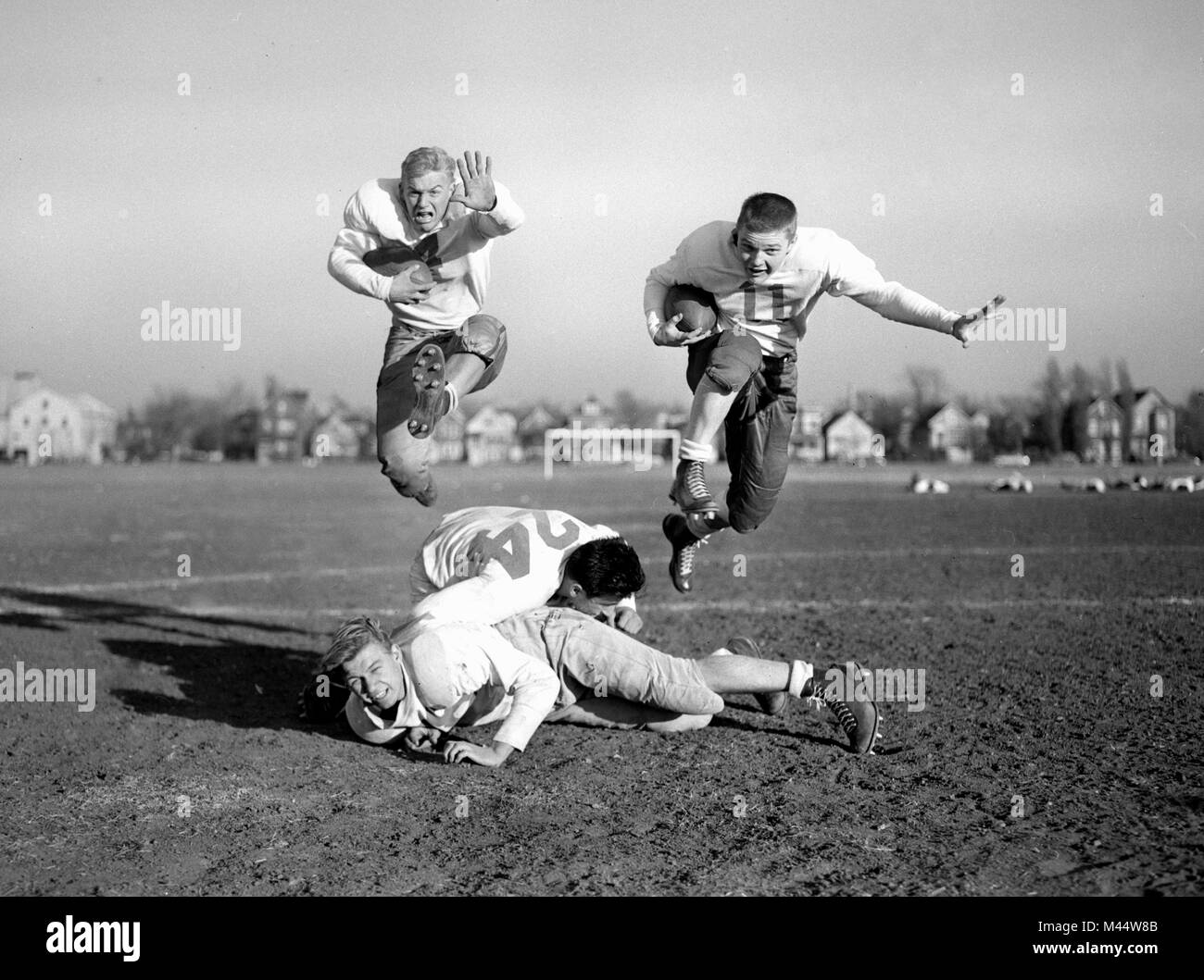 Dos futbolistas adolescentes sin cascos saltan sobre sus compañeros, replicando la acción del juego durante la práctica, ca. 1950. Foto de stock