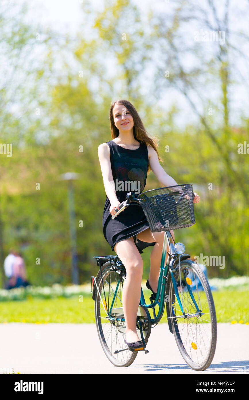Vista vertical de chica en bicicleta vistiendo el traje corto negro. Joven Mujer feliz cabalgando por carretera en el muelle verde parque al libre. Chica deportiva ri Fotografía de stock -