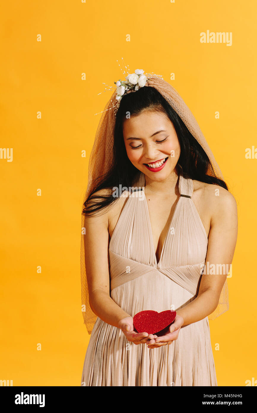 Asian bride en beige vestido de novia sosteniendo un corazón rojo aislado en naranja Foto de stock