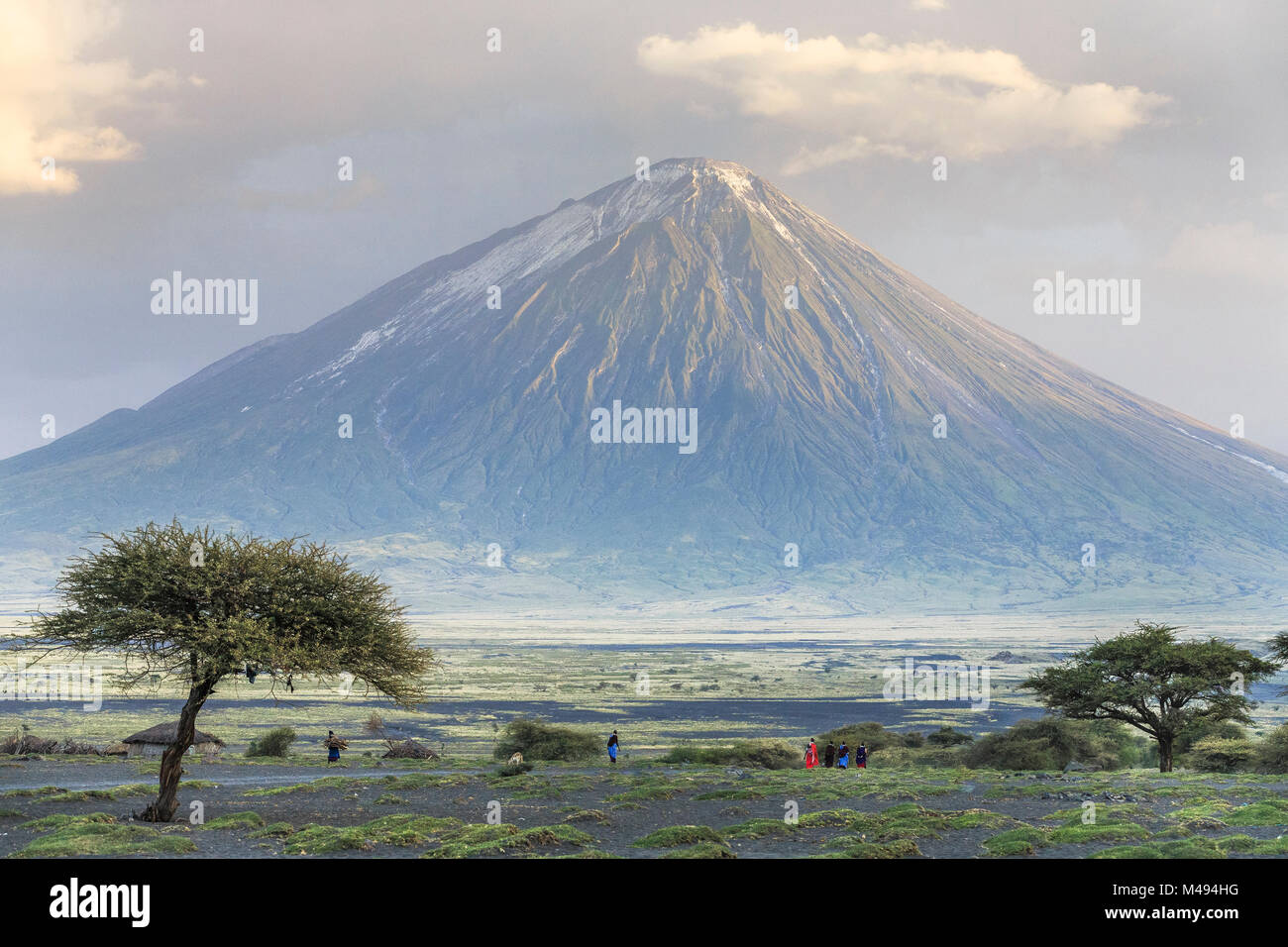 Ol Doinyo Lengai, un volcán activo celebrado en alta estima y conocida localmente como la Montaña de Dios por los Masai, con personas y cabañas en la sabana de abajo, el Valle del Rift, Tanzania Foto de stock