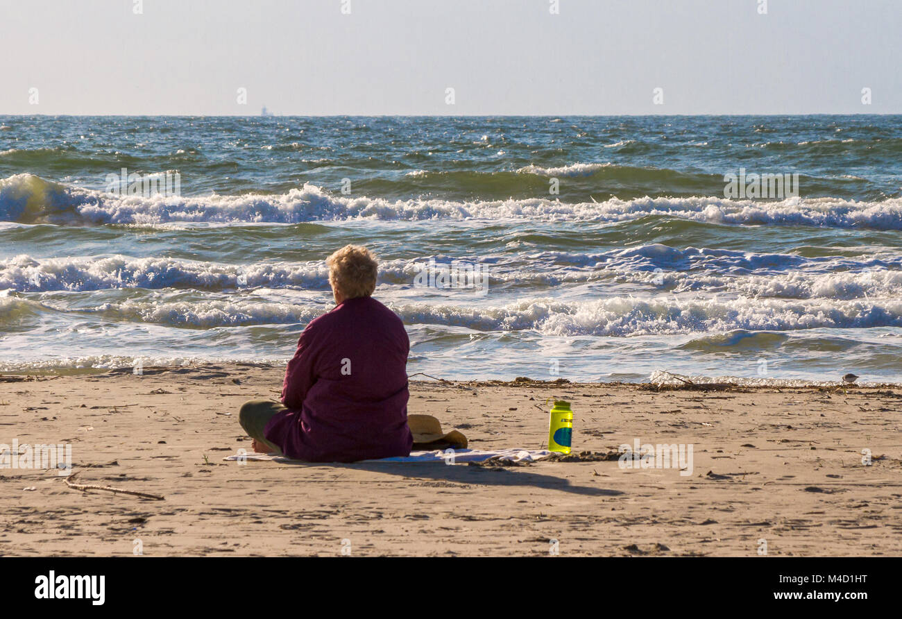 Un estadounidense senior citizen mujer sentada en la playa y mira el océano en el sol por la mañana temprano. Foto de stock