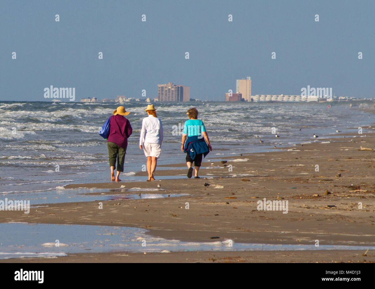 Tres mujeres senior citizen americana caminar juntos en una playa en la mañana temprano con edificios en el fondo. Foto de stock