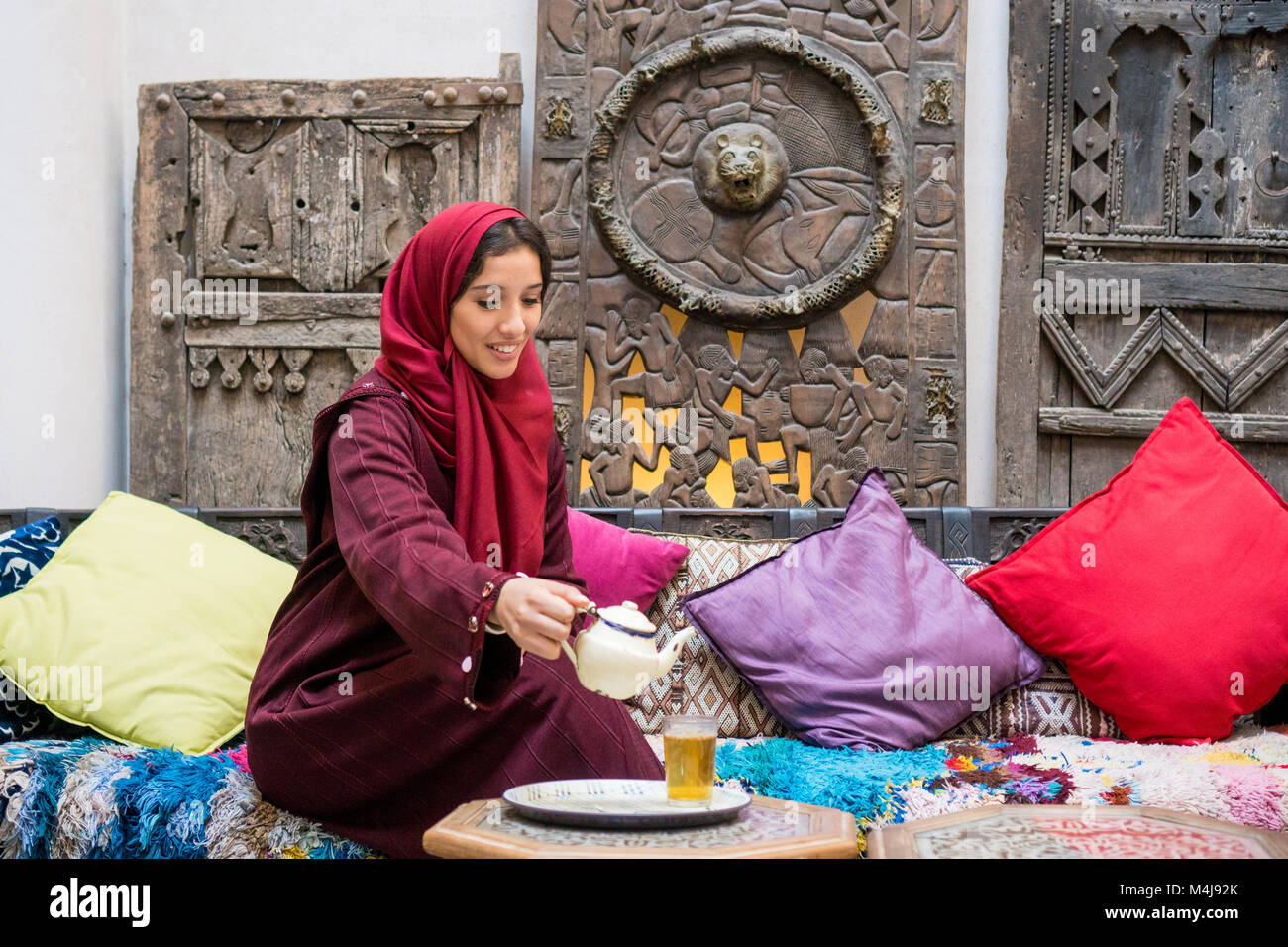 Mujer árabe en el tradicional vestido rojo con hijab derramando sobre su cabeza un té en el tradicional ambiente de oriente medio Foto de stock