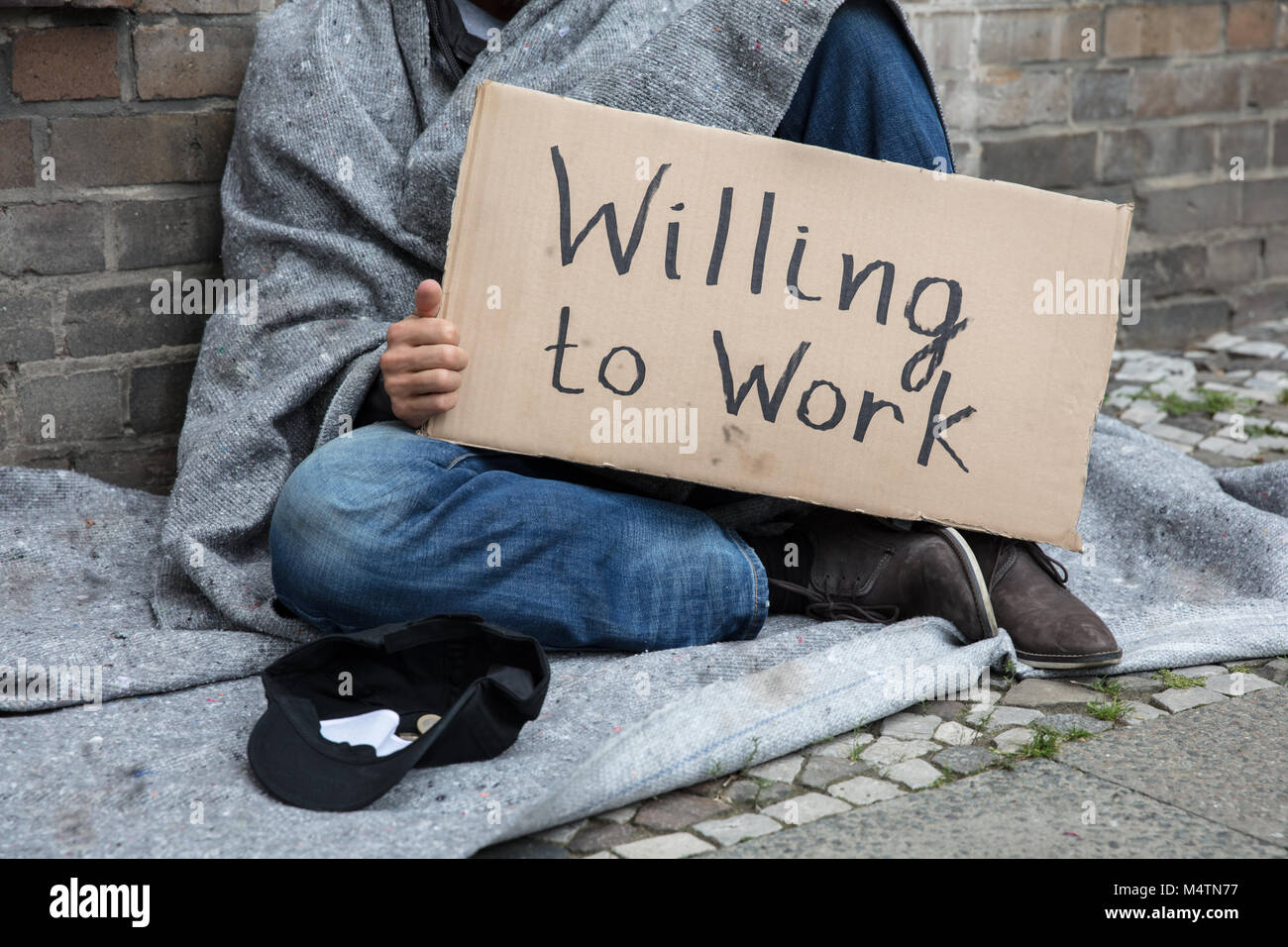 Hombre sin hogar en el capó sentados en la calle la celebración de cartón con texto dispuestos a trabajar Foto de stock