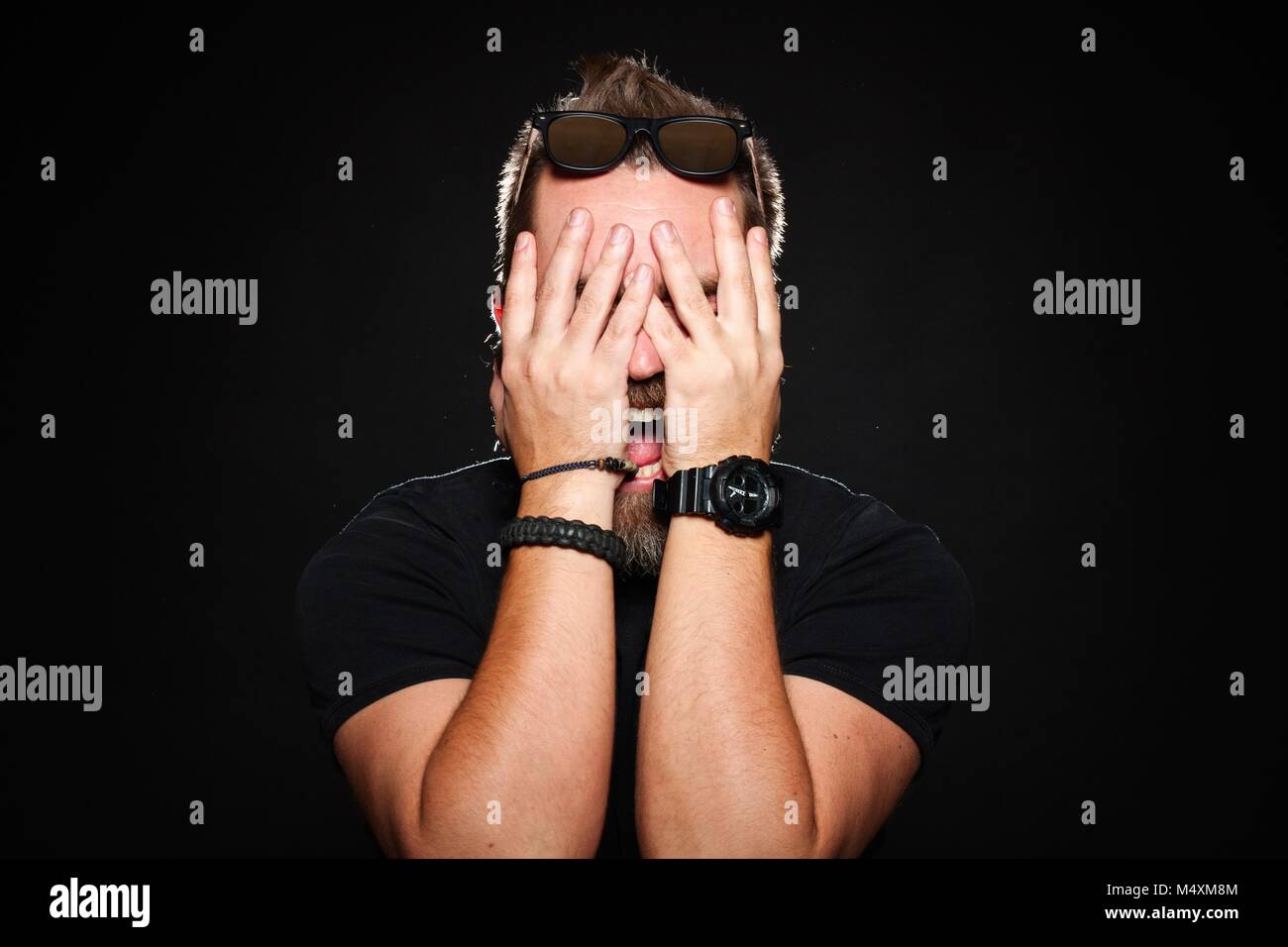Un hombre barbado mantiene sus manos detrás de su rostro y gritos en studio sobre un fondo negro Foto de stock
