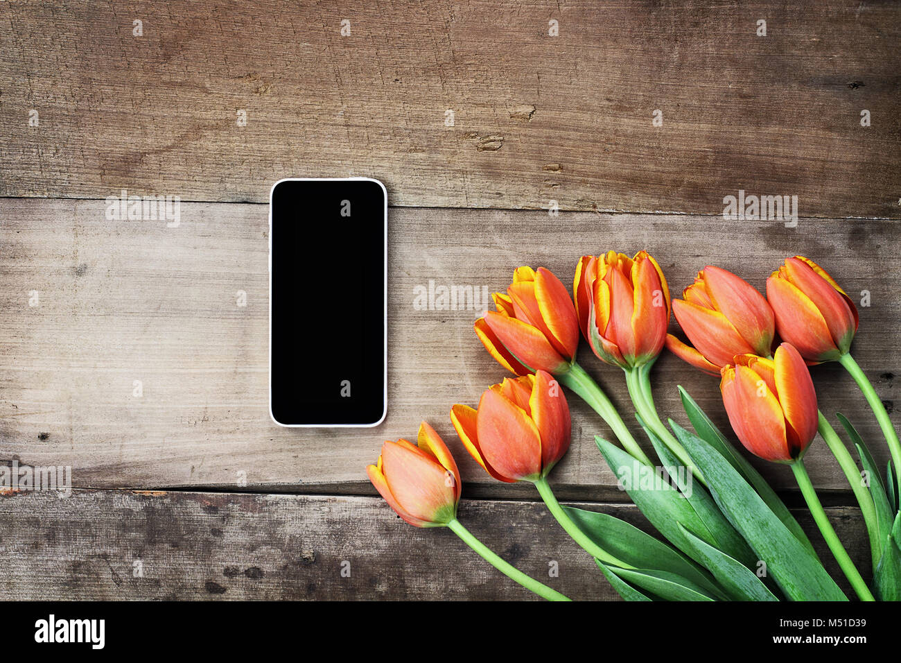 Fotografía cenital un teléfono celular en blanco con un Ramo de tulipanes naranjas y amarillas sobre una tabla de madera. Plana Vista superior laicos estilo. Foto de stock