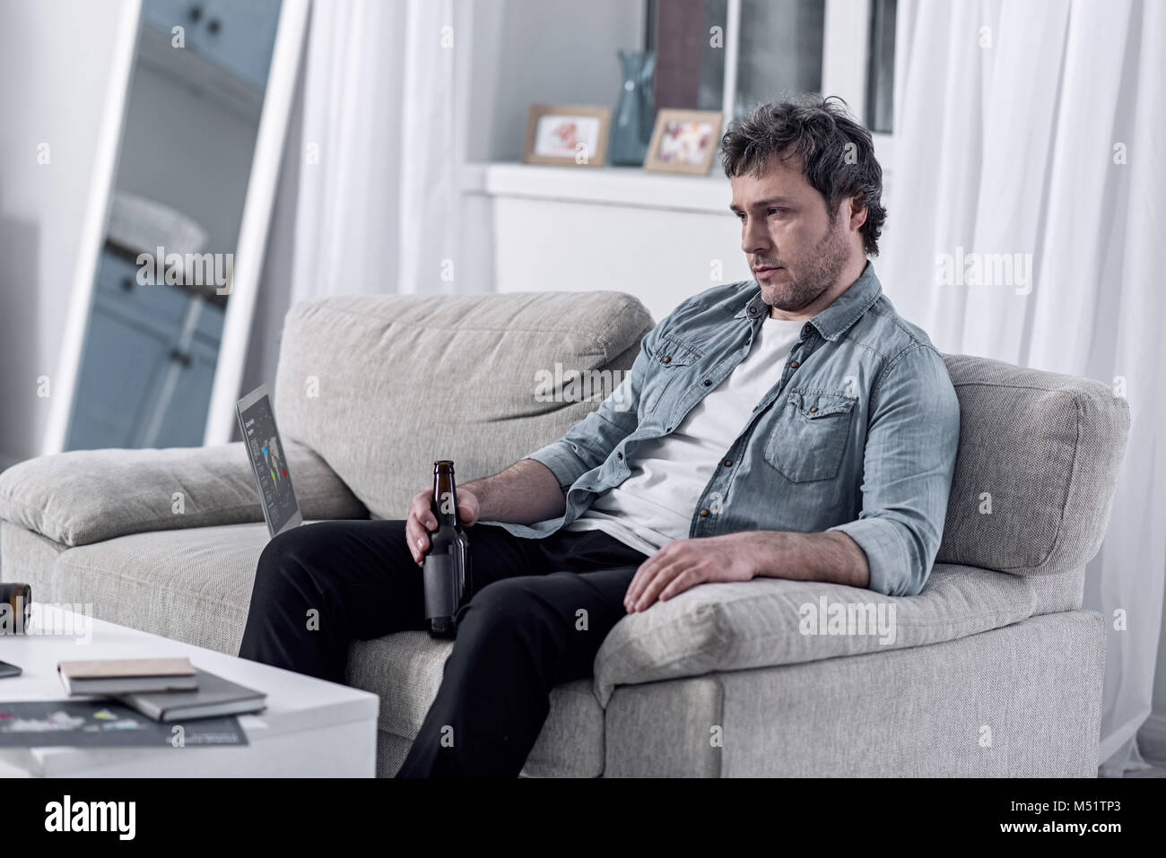 Unshaven hombre sentado en su casa y bebiendo cerveza después de perder su puesto de trabajo Foto de stock
