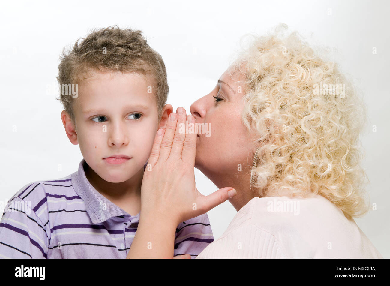 Vista lateral de la cabeza y los hombros de una mujer de pelo rizado rubio susurrando en la oreja de un niño de 8 años de edad delante de un fondo blanco. Foto de stock