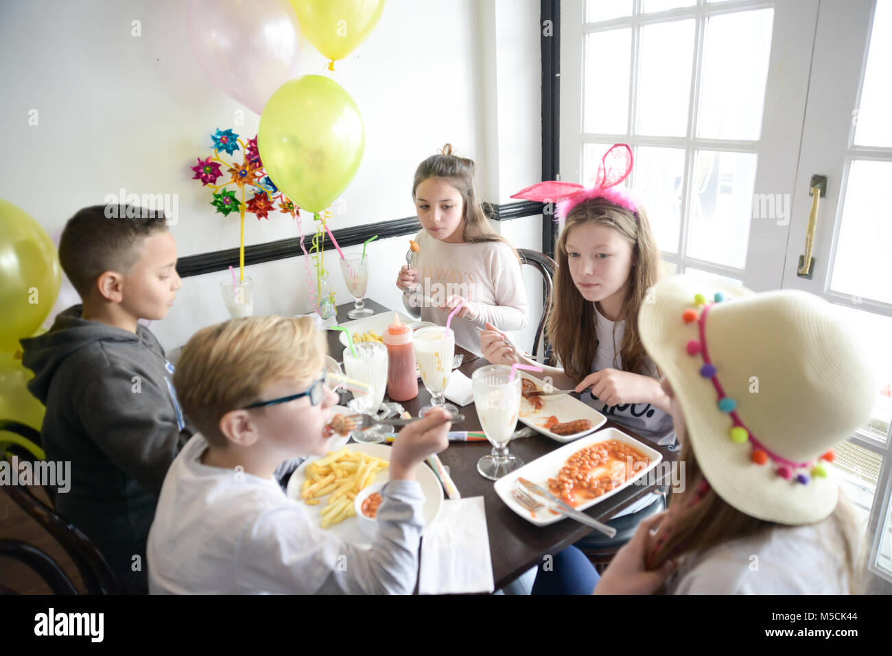 Cinco niños pequeños están sentados en una mesa comiendo parte alimentos fritos y beber batidos- hay globos y decoraciones del partido Foto de stock