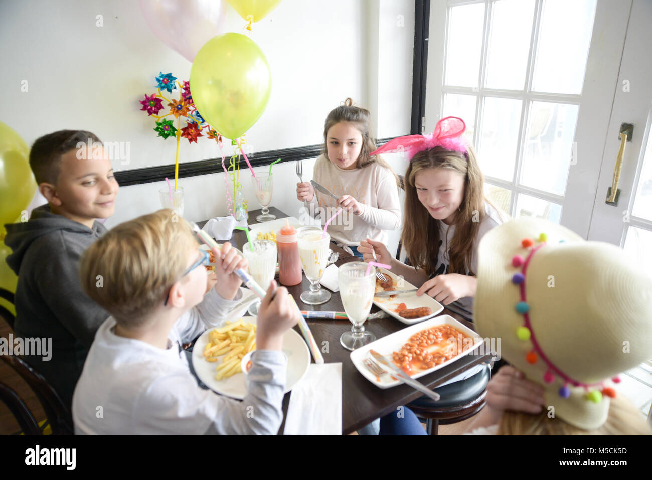 Cinco niños pequeños están sentados en una mesa comiendo parte alimentos fritos y beber batidos- hay globos y decoraciones del partido Foto de stock