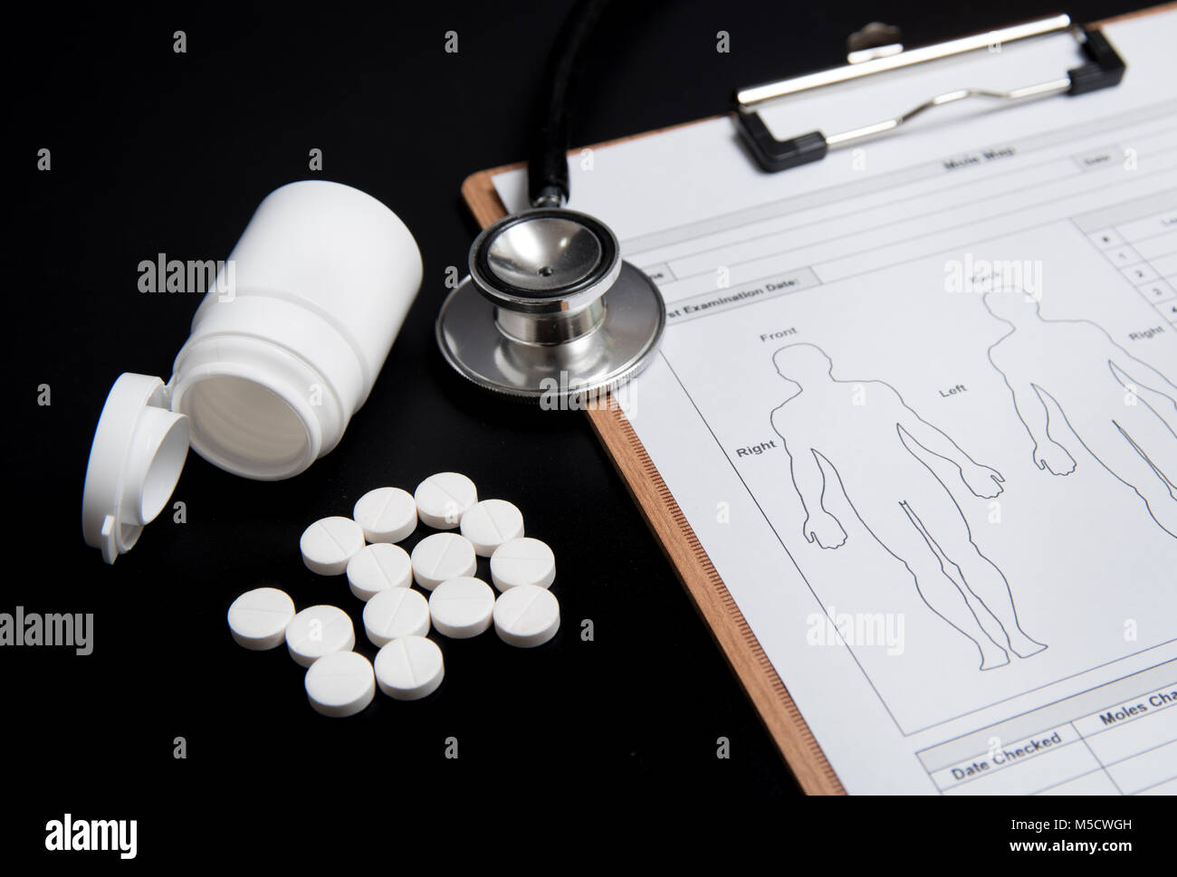 Pastillas de color blanco y una botella blanca, junto con un estetoscopio y una ficha médica, están sobre un fondo negro. Foto de stock
