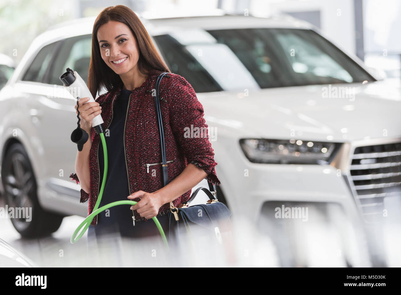 Retrato sonriente, seguros de mujer sosteniendo el cable de carga del híbrido en concesionario de coches showroom Foto de stock