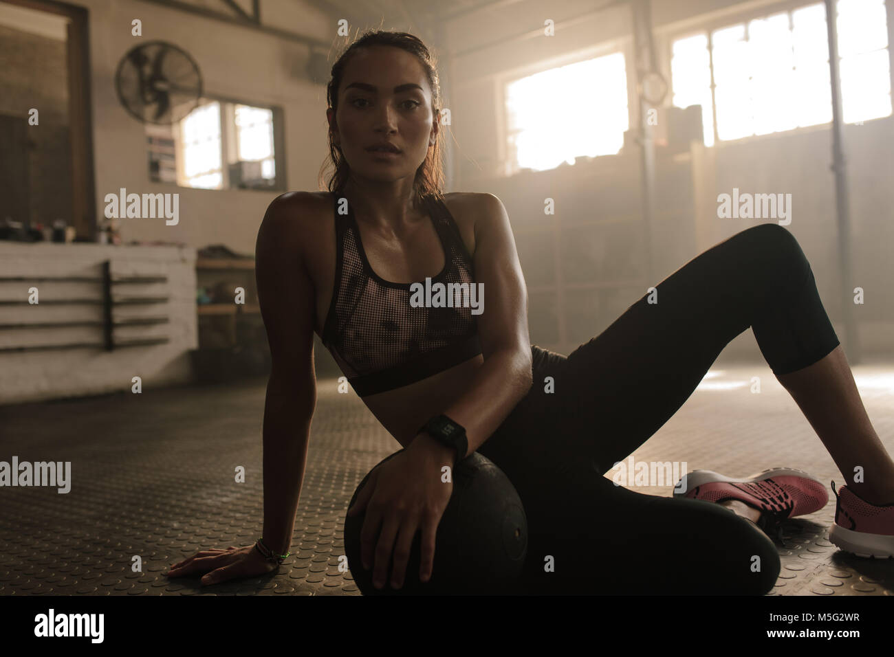 Mujer joven sentado en el piso del gimnasio con balón medicinal tras la sesión de ejercicio. Mujeres tomando un descanso del entrenamiento. Foto de stock