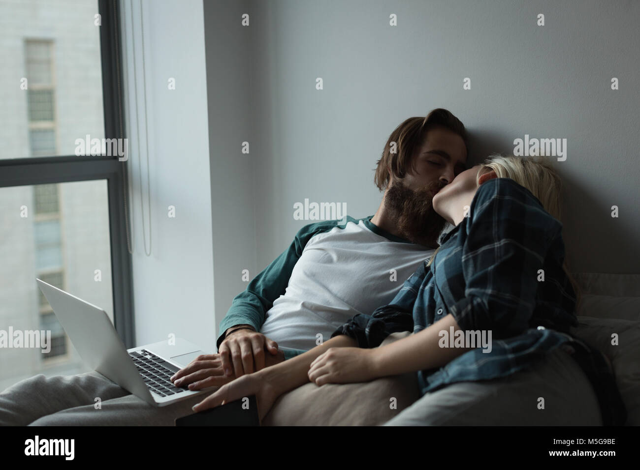 Pareja besándose mutuamente mientras usa portátil en casa Foto de stock