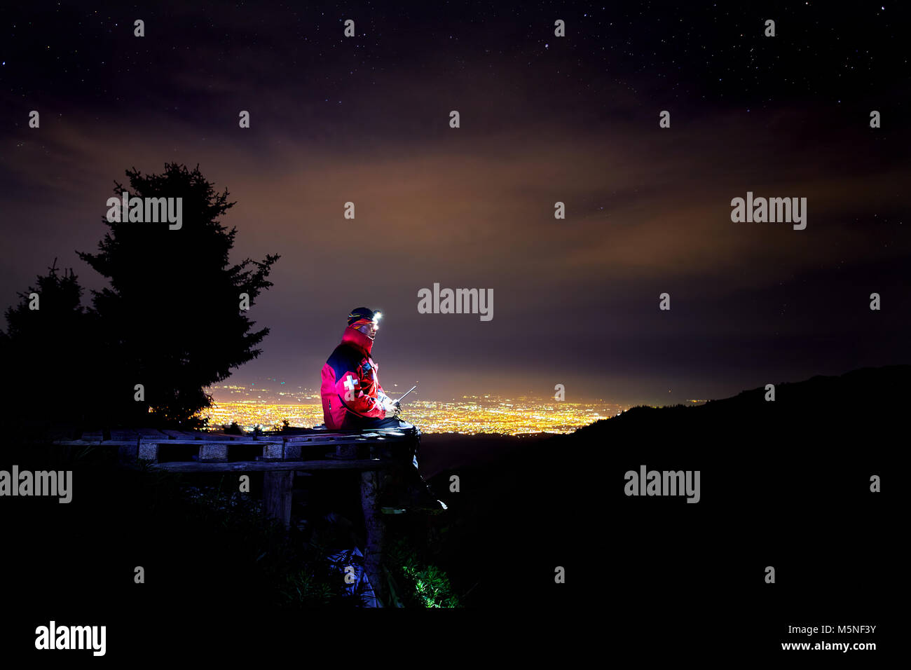 Guardavidas de chaqueta roja con cruz médica el emplazamiento en la rama de madera en la estación de esquí de montaña con vistas a la ciudad de noche la luz Foto de stock