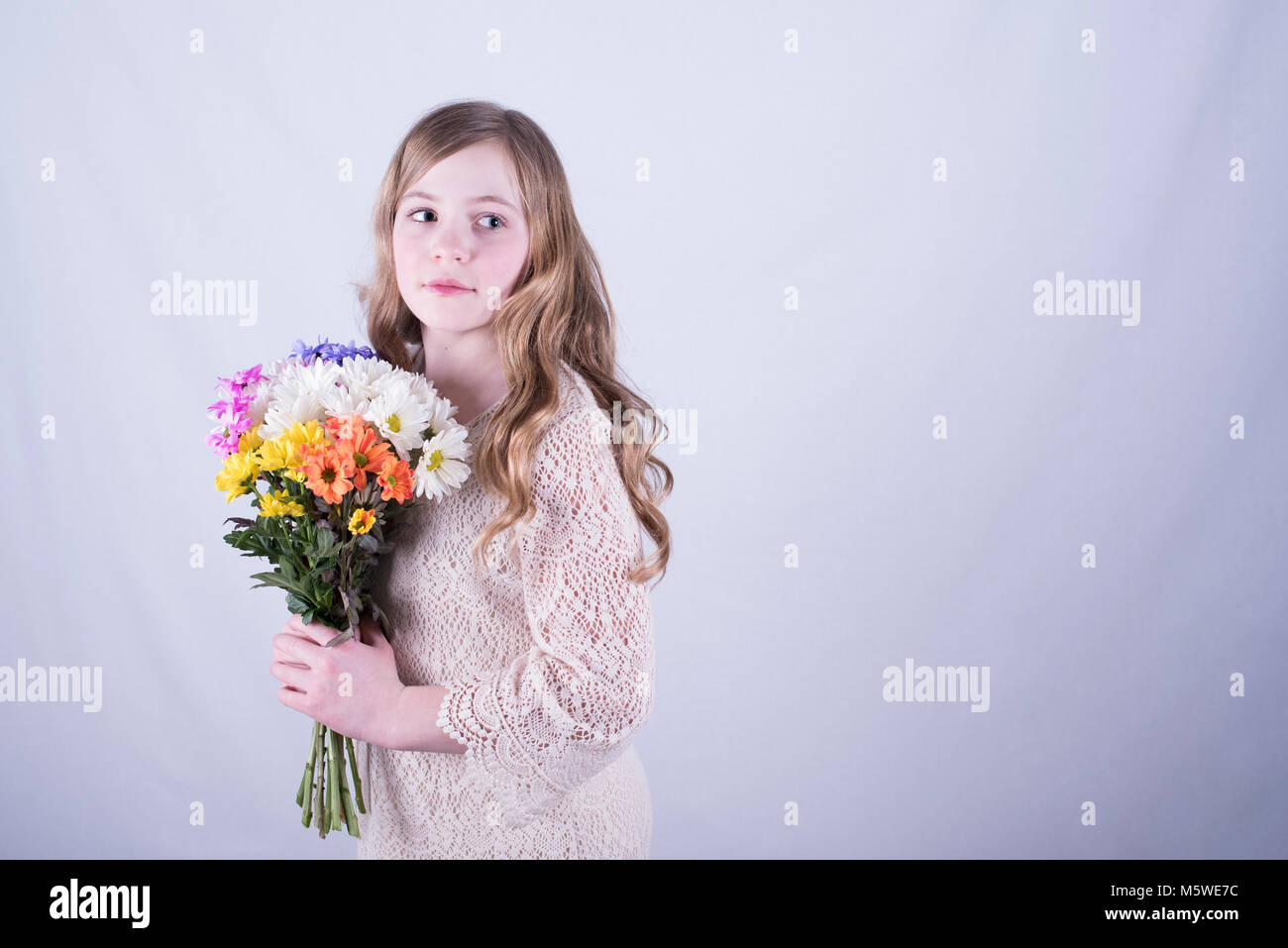12-year-old girl con largo pelo rubio sucio, girada y celebración colorido bouquet de margaritas mientras mirando hacia el lado, fondo blanco. Foto de stock