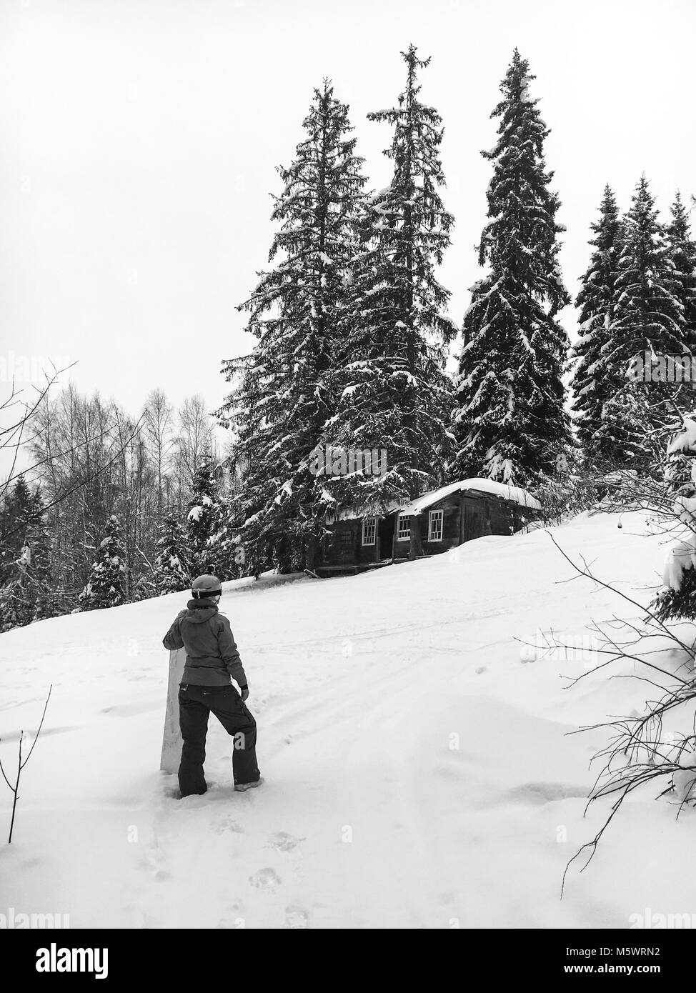 Una chica de snowboard en un casco azul destaca con su mano sobre el lado de una placa de color naranja brillante en el bosque y se centra en una antigua casa de madera abandonada h Foto de stock
