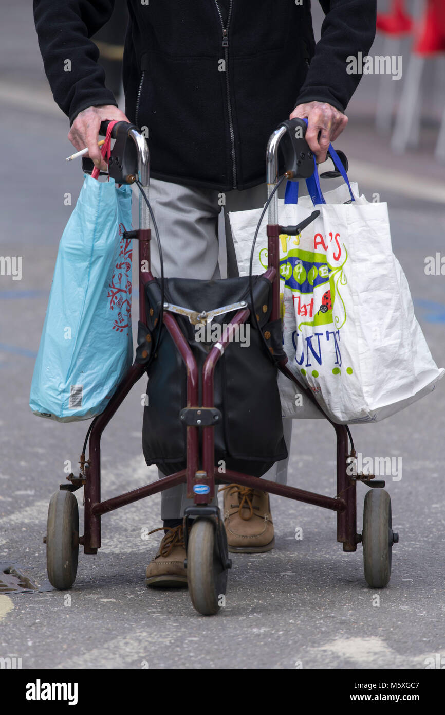 Un viejo pensionista utilizando un bastidor de soporte mientras zimmer caminando por la calle llevando bolsas de compras en Cardiff, Gales, Reino Unido. Foto de stock