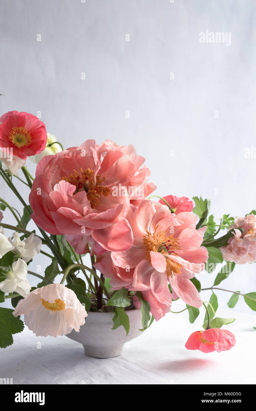 Cerca de peonías: arreglo floral con arrecifes de peonías, amapolas de Islandia y de las existencias, en un cuenco de cerámica blanca, sobre una mesa, con el telón de fondo de color gris pálido Foto de stock