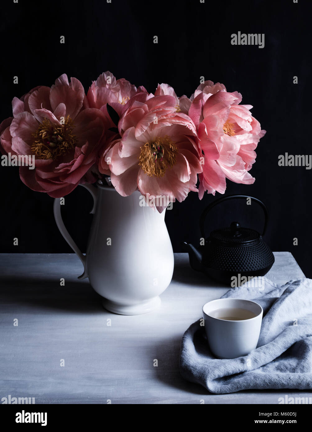 Encanto Coral peonías en flor, en una jarra blanca sobre la mesa con taza y tetera tetsublin, fondo oscuro, rodada en luz natural Foto de stock