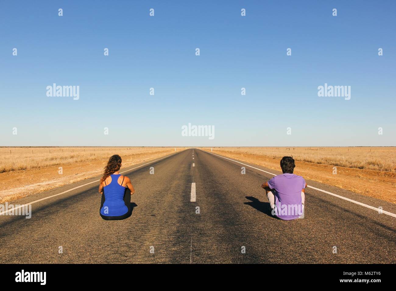 Pareja joven desde detrás sentada en una interminable recta carretera vacía en el medio de la nada en el Outback de Australia. Los mochileros, concepto visionario Foto de stock
