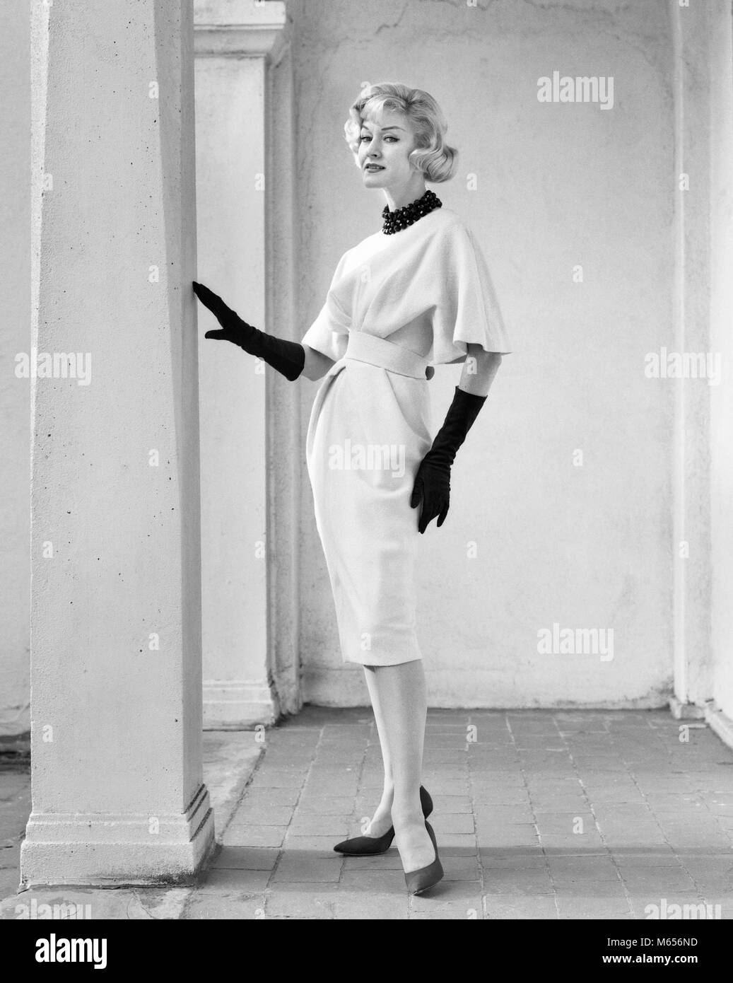 1960 Retrato de longitud completa elegante mujer rubia vistiendo ropa con  mangas DRAPEADAS COMPLETO GUANTES largos posando POR COLUMNA AL AIRE LIBRE  - F11349 DEB001 HARS VESTIDAS NOSTALGIA contacto ocular 20-25 AÑOS