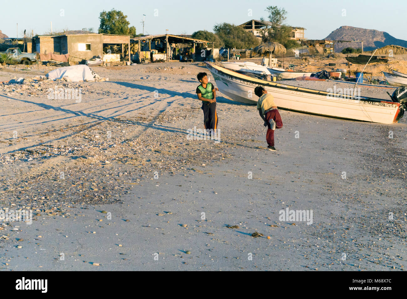 Dos pequeños niños juegan mexicanos arrojando piedras al mar desde la playa donde barcos de pesca están hacia arriba y la costa está bordeada de casas improvisadas de pescadores Foto de stock