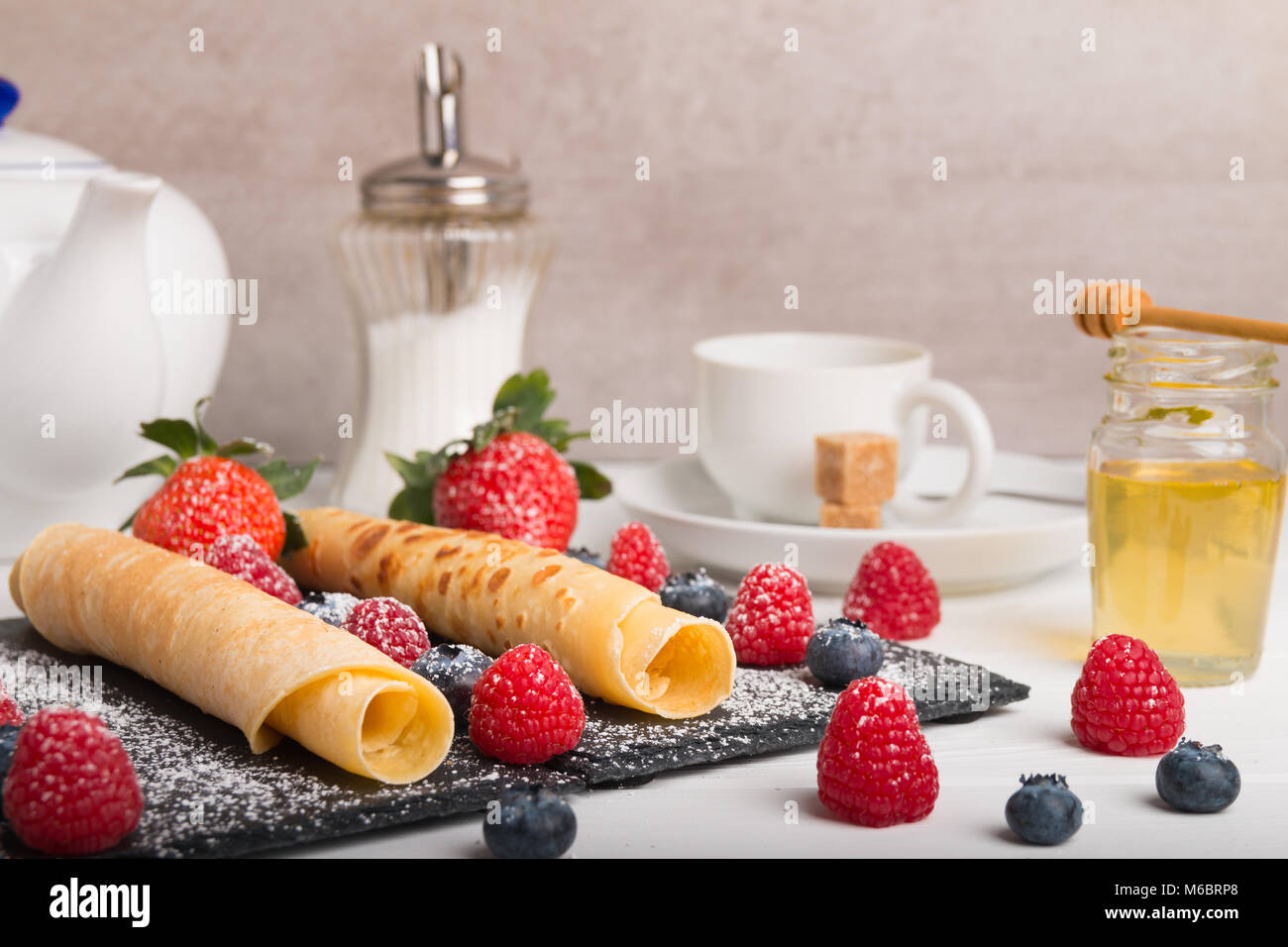 Rodó crepes con fresas frescas y de frambuesa y arándanos, sobre una placa de piedra negra sobre una tabla de madera blanca con miel y azúcar blanco y Foto de stock