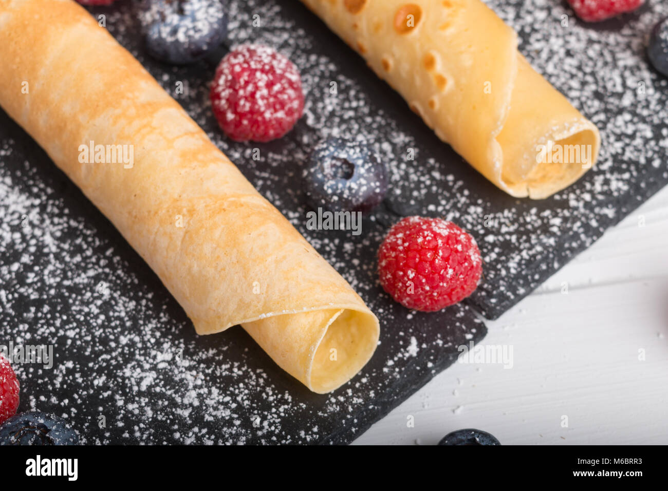 Rodó crepes con fresas frescas y de frambuesa y arándanos, sobre una placa de piedra negra sobre una tabla de madera blanca con miel y azúcar blanco y Foto de stock