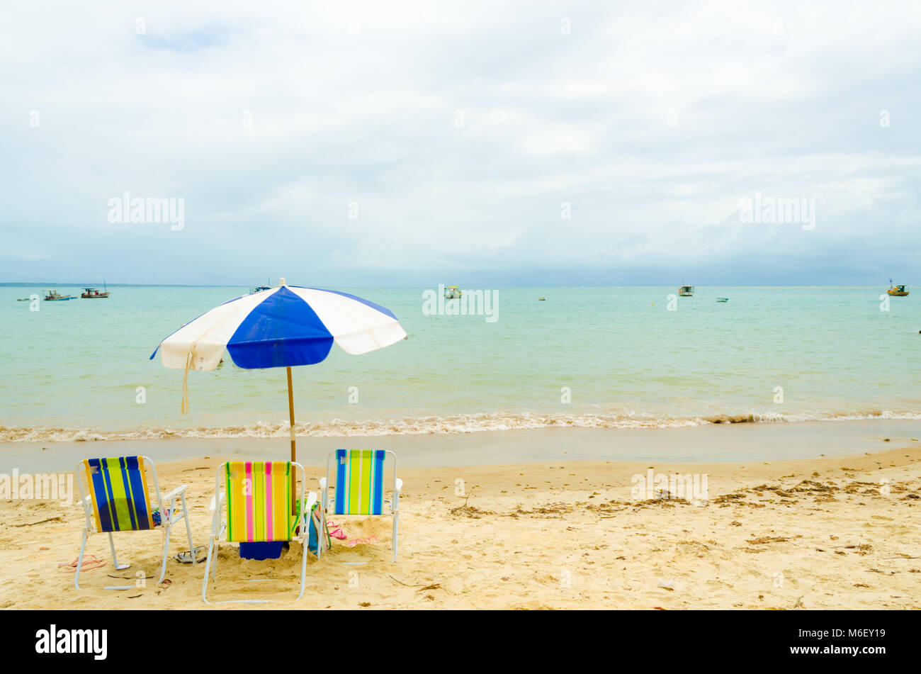 Playa aislada con tres coloridos sillas de playa y un paraguas de color azul y blanco. Una pequeña ola rompe en la arena y algunos barcos están en el mar. Foto de stock