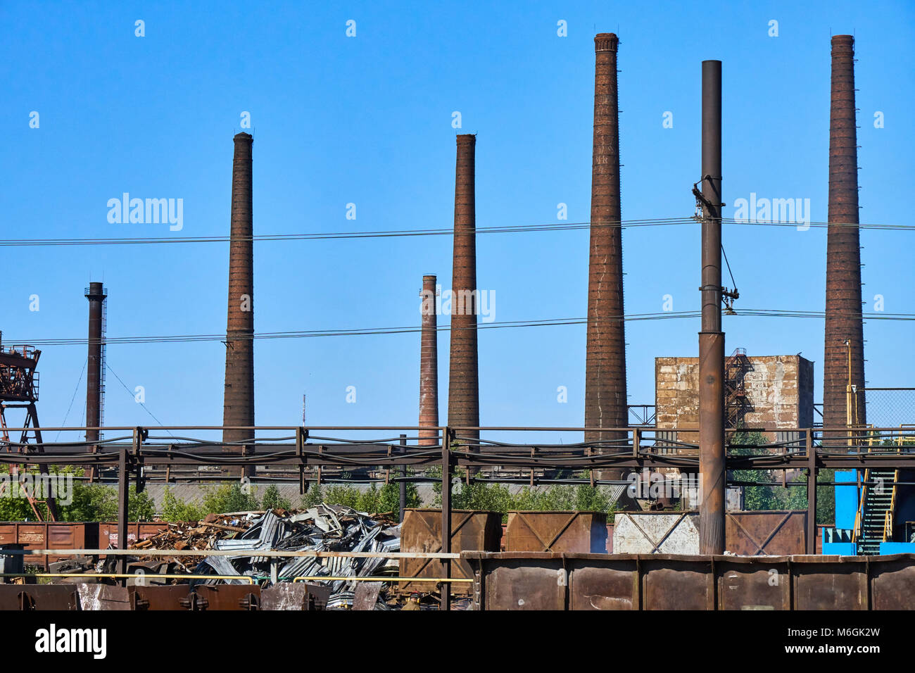 Complejo industrial abandonado, con estacas de humo altas y desgastadas que se elevan sobre los edificios en ruinas contra un cielo azul claro Foto de stock