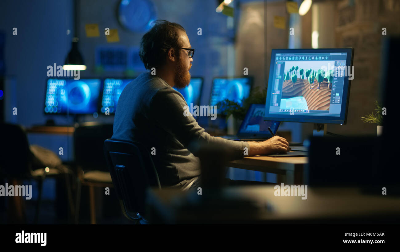 Desarrollador de Aplicaciones Móviles macho trabaja con gráficos en su PC con dos monitores. Trabaja a altas horas de la noche, en una oficina vacía. Foto de stock