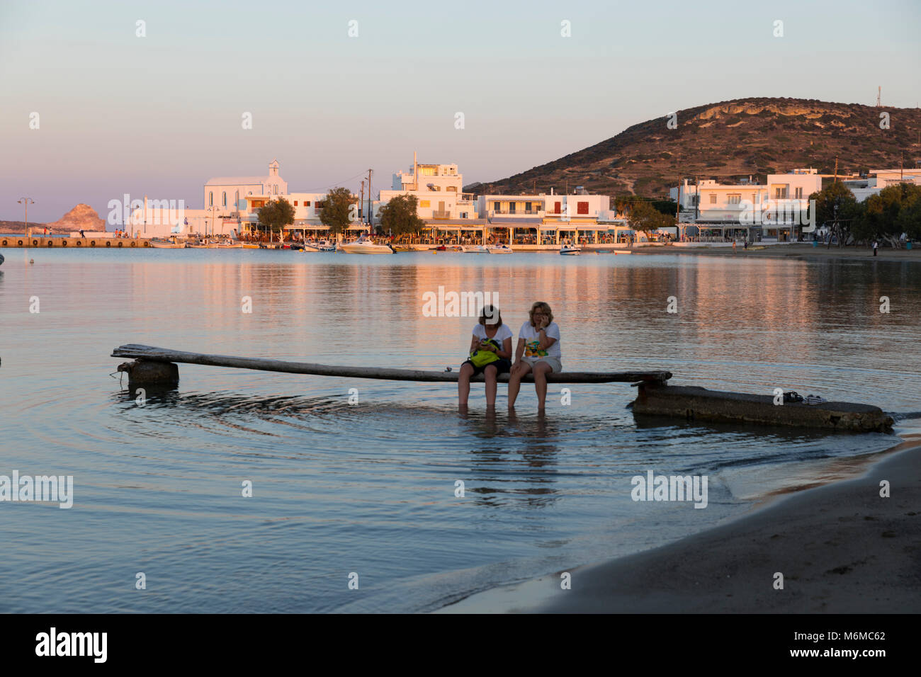 Vista a lo largo de la playa con 2 mujeres de sat en el muelle al atardecer, Pollonia, Milos, Cyclades, islas griegas del Mar Egeo, Grecia, Europa Foto de stock