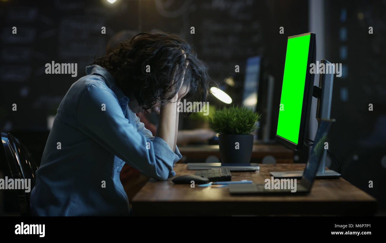 Estresado, sobrecargado de trabajo Diseñador femenino sostiene a su cabeza entre las manos mientras se trabaja en un ordenador personal con pantalla verde Croma Key. Foto de stock