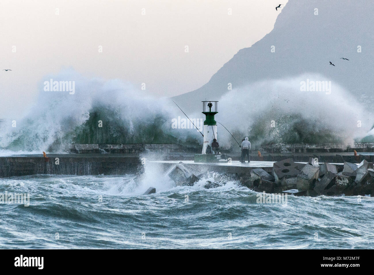 Los pescadores sorteando enormes olas que pueden verse aquí reventándose contra el rompeolas de Kalk Bay Harbour en Cape Town, Sudáfrica. Foto de stock