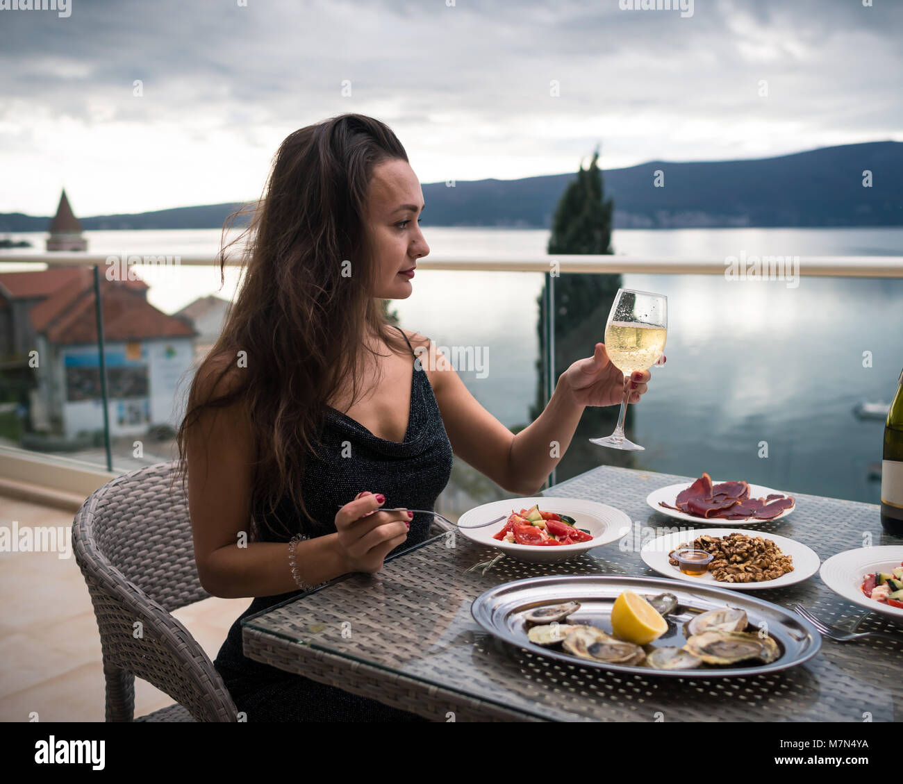 Joven Mujer sentada en un balcón en la mesa con la comida local y sostiene una copa de vino espumoso. Chica está mirando el mar y las montañas. Vista lateral Foto de stock