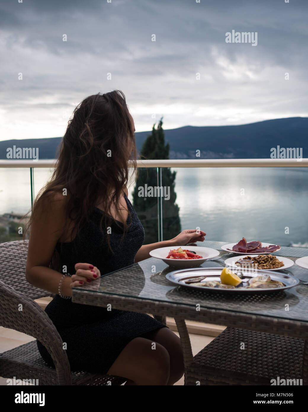 Irreconocible mujer sentada a la mesa en el balcón con ostras, carne y ensaladas. Chica está mirando el mar y las montañas. Vista lateral. Foto de stock