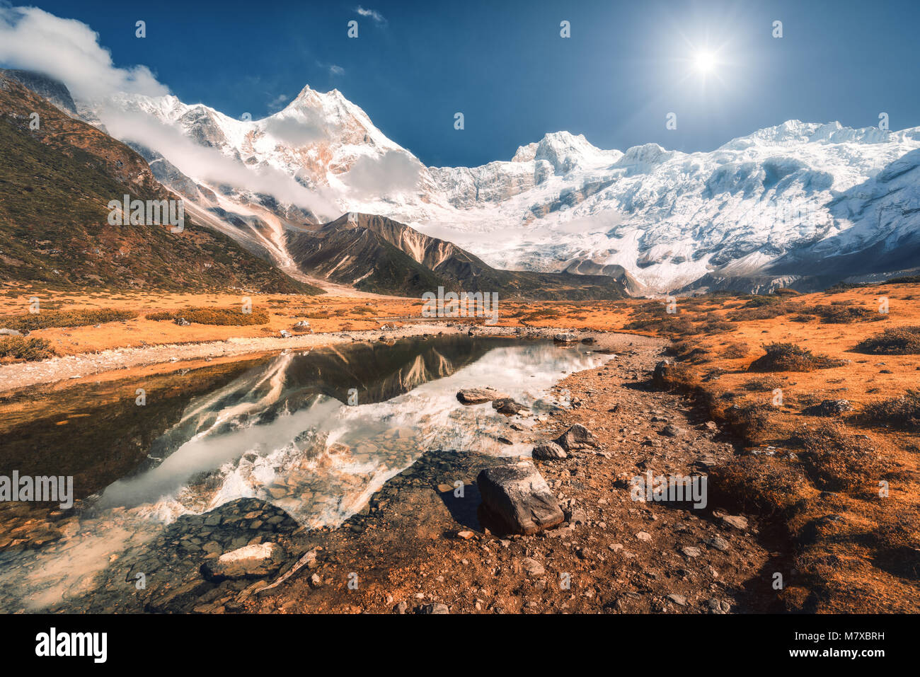 Hermosa escena con altas rocas con picos cubiertos de nieve, piedras en Mountain Lake, reflejo en el agua, el cielo azul en el atardecer. Nepal. Un paisaje impresionante wi Foto de stock