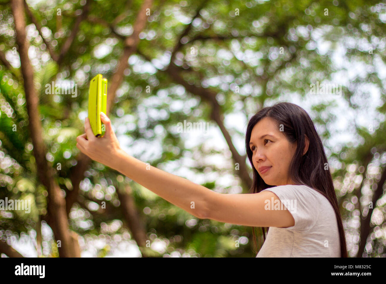 Sonriente y atractiva mujer asiática con un teléfono móvil en un parque verde, teniendo selfie Foto de stock