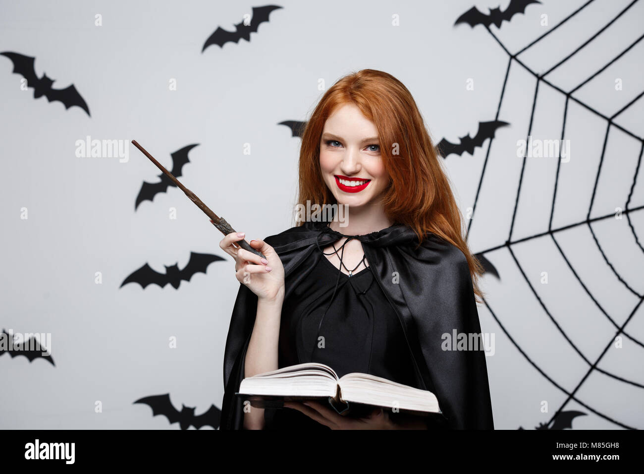 Concepto de Halloween - Hermosa Bruja jugando con la varita mágica y la magia del libro sobre fondo gris. Foto de stock