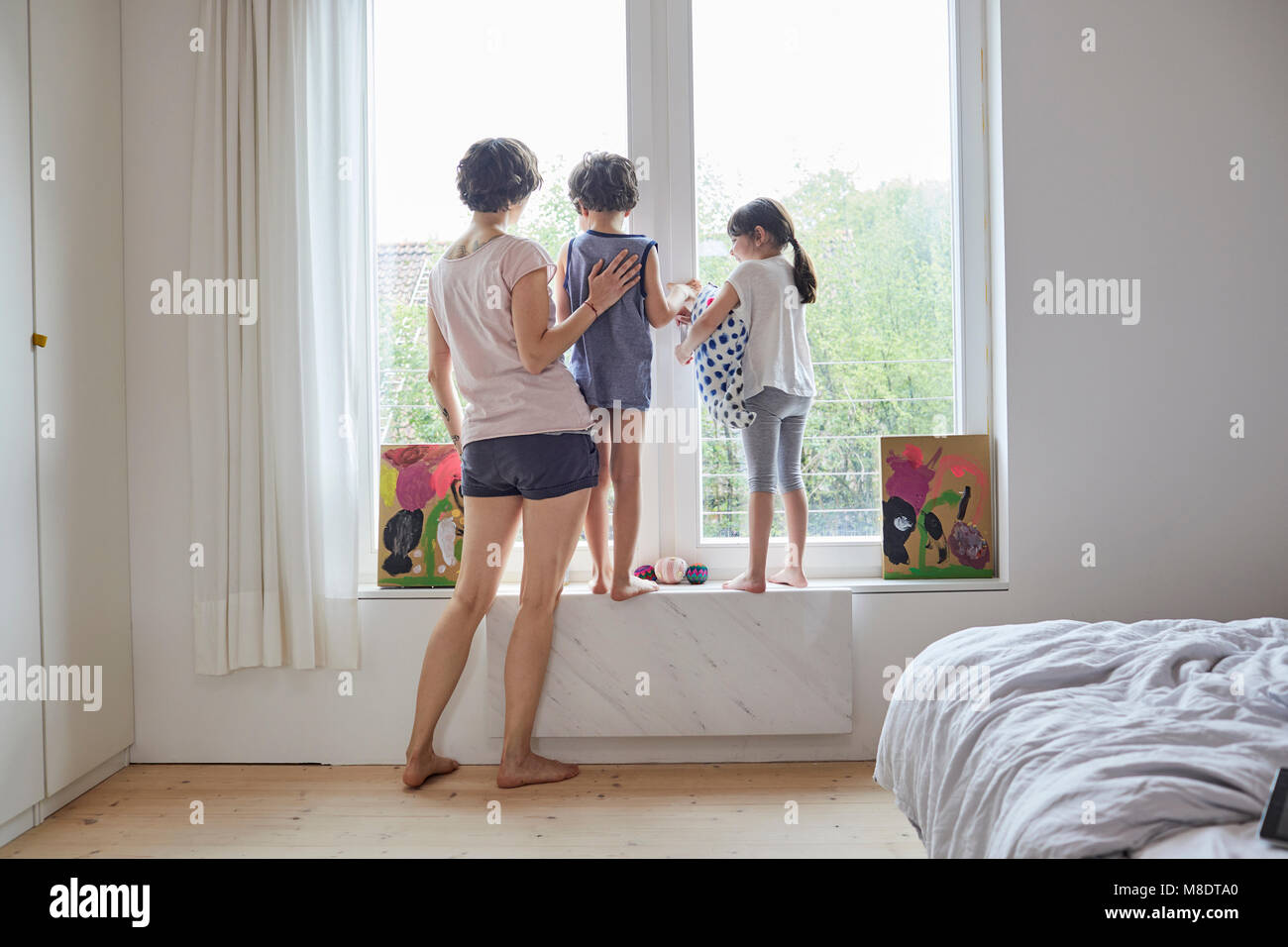Madre, hijo e hija mirando afuera de la ventana de la habitación, vista trasera Foto de stock