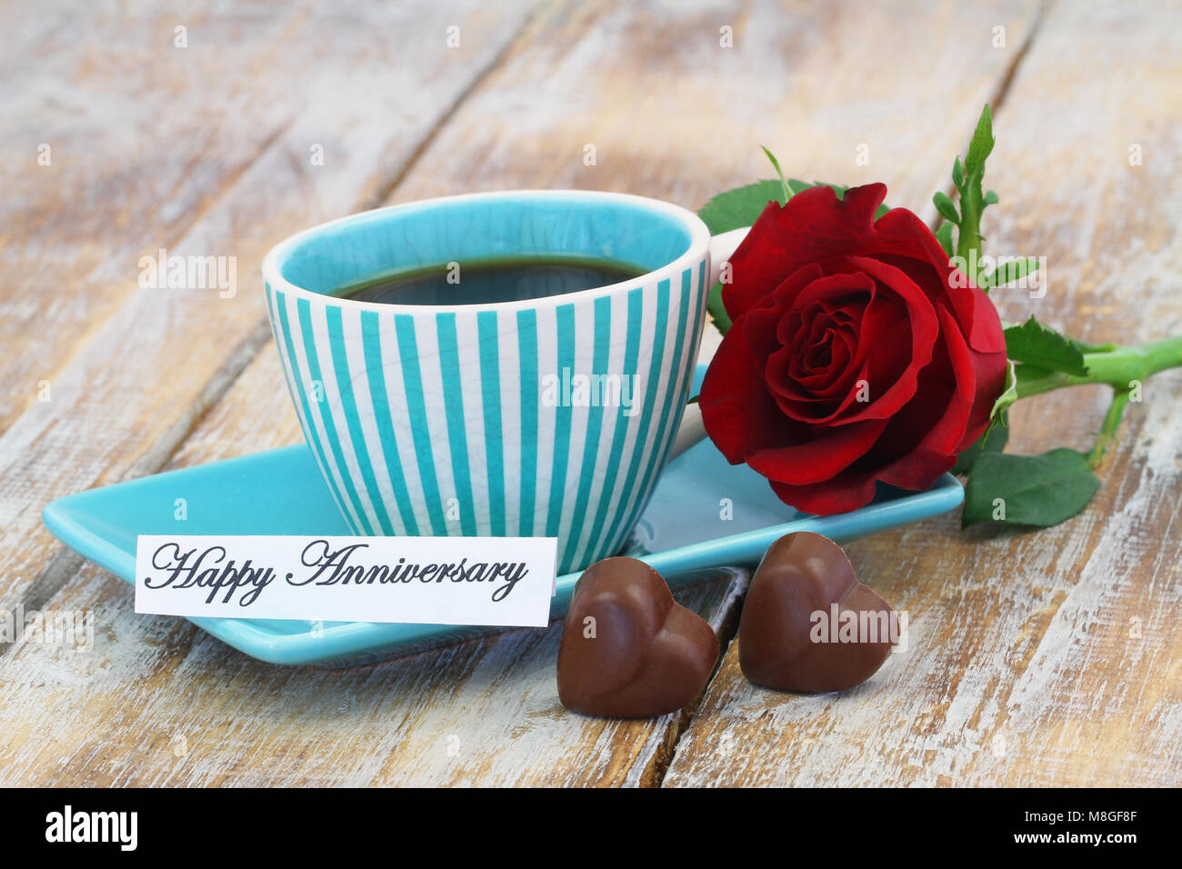 Feliz Aniversario de tarjeta con una taza de café, dos chocolates en forma de corazón y rosa roja Foto de stock