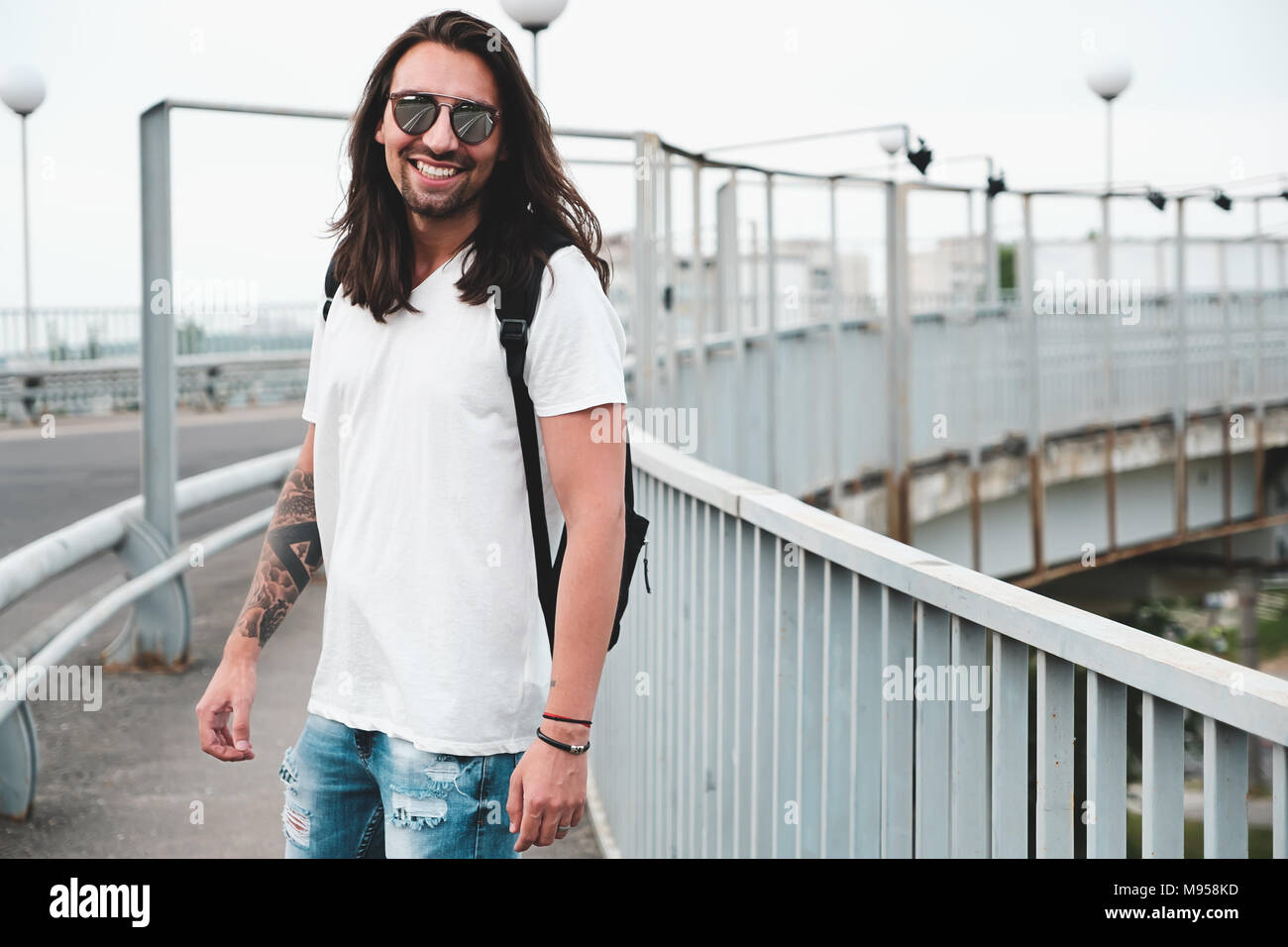 Hombre sonriente hang out, caminar por el puente urbano en la ciudad.  Alegre macho con pelo largo y gafas de sol, ropa casual es feliz de vivir  un buen día Fotografía de