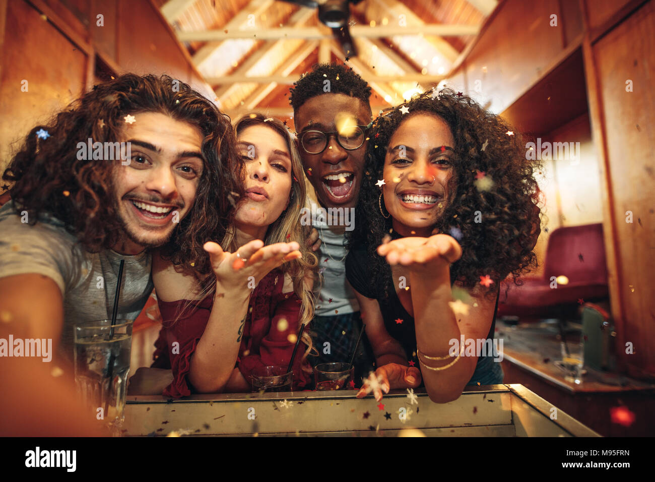 Grupo de amigos disfrutando de un partido y soplando confetti. Hombres y mujeres capturar la diversión en selfie en discoteca. Foto de stock