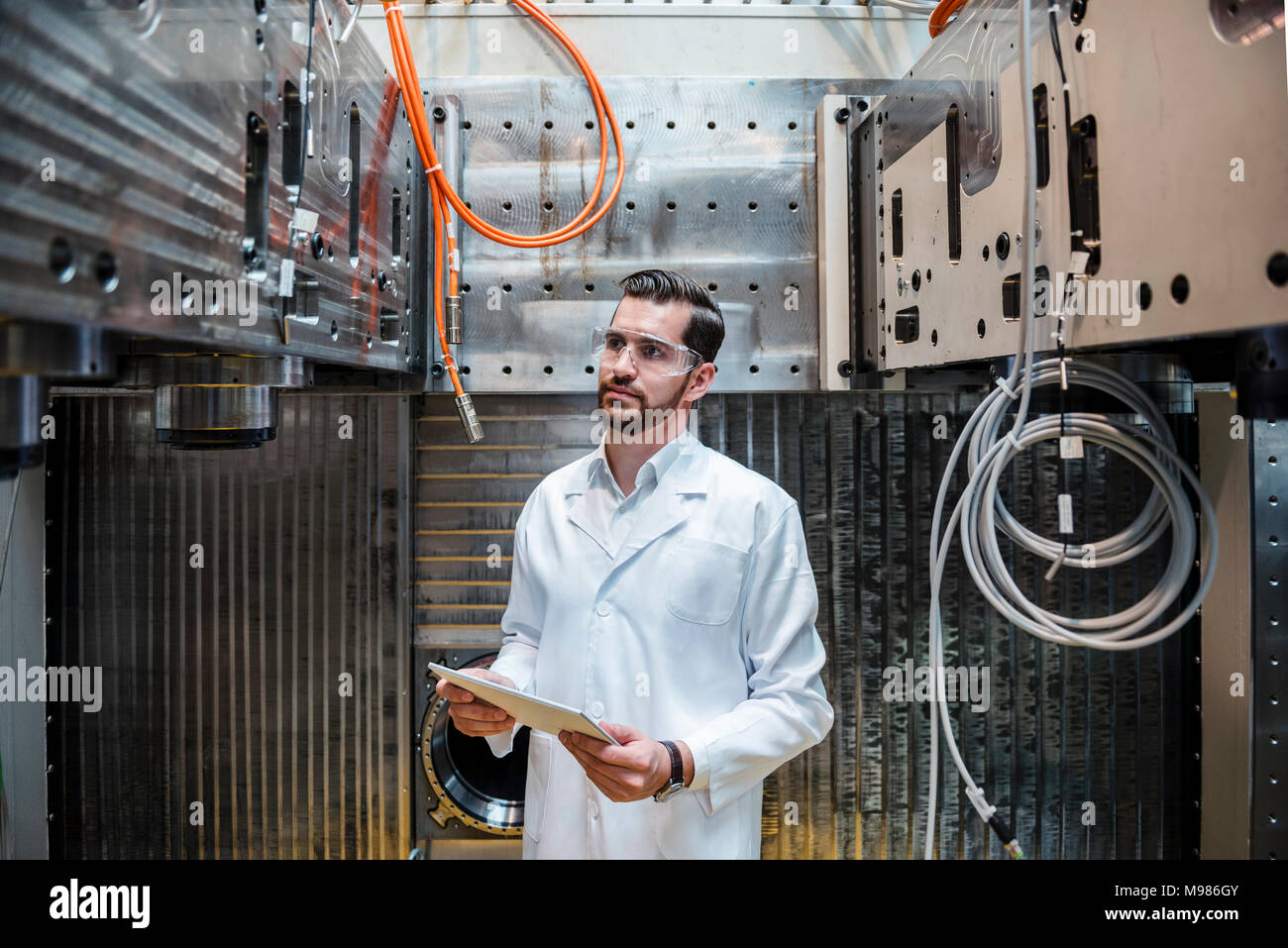 Hombre vestido con bata de laboratorio y gafas de seguridad cuando la máquina se mantiene tablet Foto de stock